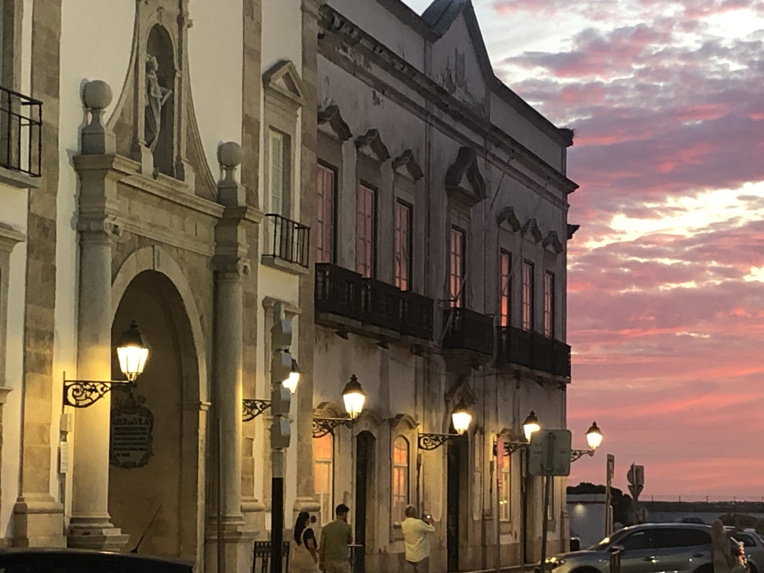 Historic buildings lit by street lamps at sunset with a colorful sky.